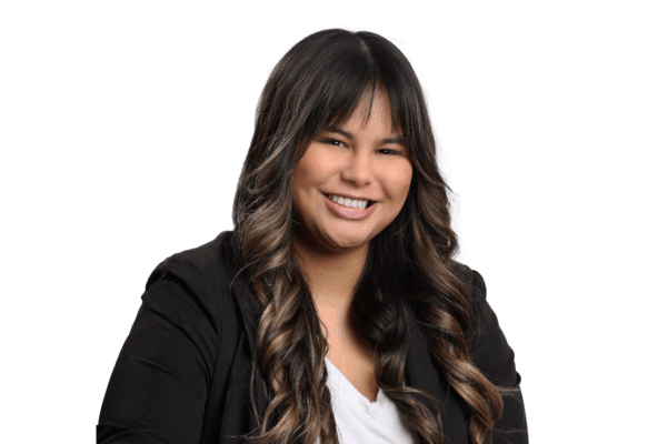 Woman with long, curled dark brown hair and bangs, wearing a black blazer and white blouse, smiling brightly in front of a white background.