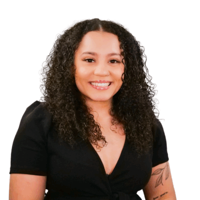 Woman with medium-length curly hair, wearing a black top and showing tattoos on her arm, smiling warmly in front of a white background.