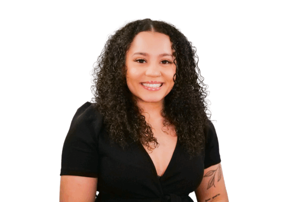 Woman with medium-length curly hair, wearing a black top and showing tattoos on her arm, smiling warmly in front of a white background.