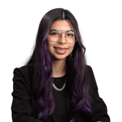 Woman with long dark hair dyed purple at the ends, wearing glasses, a black top, and a silver necklace, smiling gently in front of a white background.