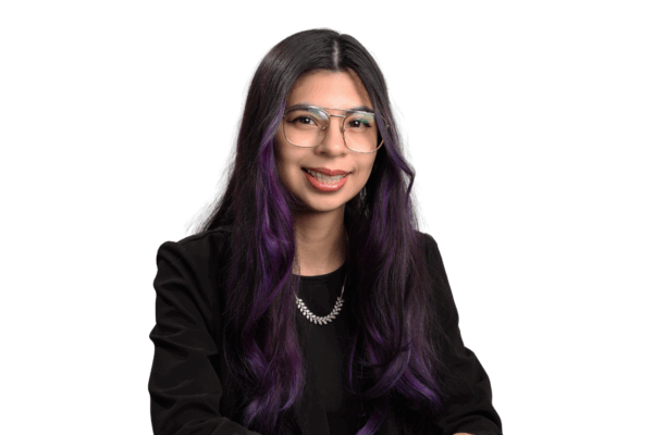 Woman with long dark hair dyed purple at the ends, wearing glasses, a black top, and a silver necklace, smiling gently in front of a white background.