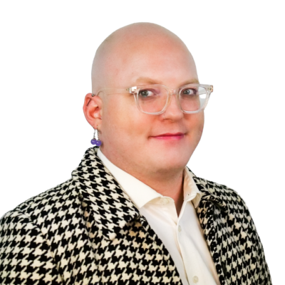 Bald person with clear glasses and a bold earring, wearing a black and white houndstooth jacket over a white shirt, smiling slightly.