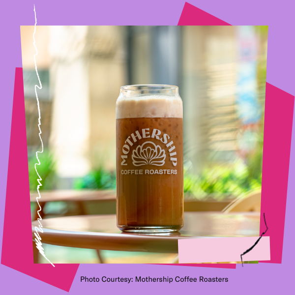 A glass of iced coffee with foam on top sits on a round outdoor table. The glass is labeled “Mothership Coffee Roasters” with a seashell logo. The background is softly blurred, showing greenery and a sunlit urban setting. The image is framed with playful pink and purple geometric shapes. Text at the bottom reads: “Photo Courtesy: Mothership Coffee Roasters.”