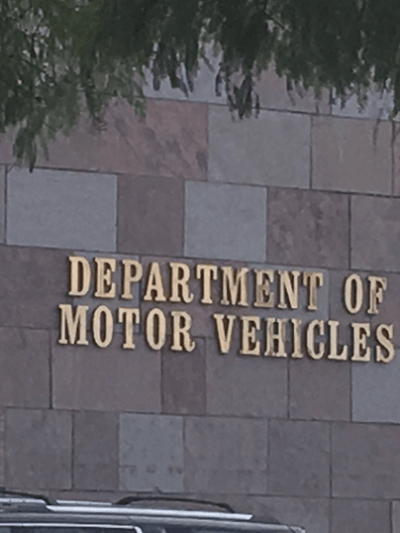 Photo of the exterior of a Department of Motor Vehicles (DMV) building. The wall is made of square and rectangular stone tiles in shades of beige and brown. Mounted in large gold letters is the text “DEPARTMENT OF MOTOR VEHICLES.” A portion of a parked vehicle and tree branches are visible in the foreground.