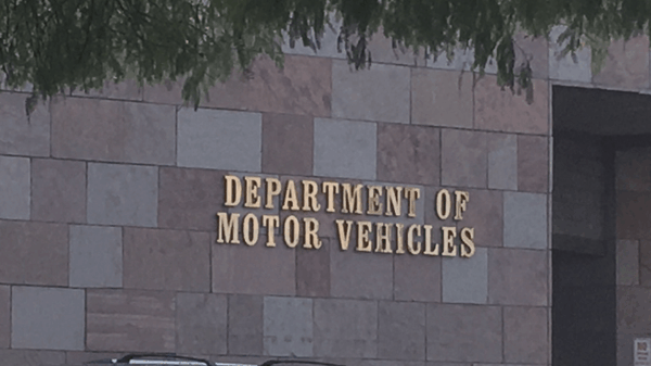 Photo of the exterior of a Department of Motor Vehicles (DMV) building. The wall is made of square and rectangular stone tiles in shades of beige and brown. Mounted in large gold letters is the text “DEPARTMENT OF MOTOR VEHICLES.” A portion of a parked vehicle and tree branches are visible in the foreground.