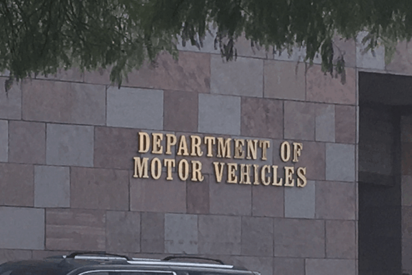 Photo of the exterior of a Department of Motor Vehicles (DMV) building. The wall is made of square and rectangular stone tiles in shades of beige and brown. Mounted in large gold letters is the text “DEPARTMENT OF MOTOR VEHICLES.” A portion of a parked vehicle and tree branches are visible in the foreground.