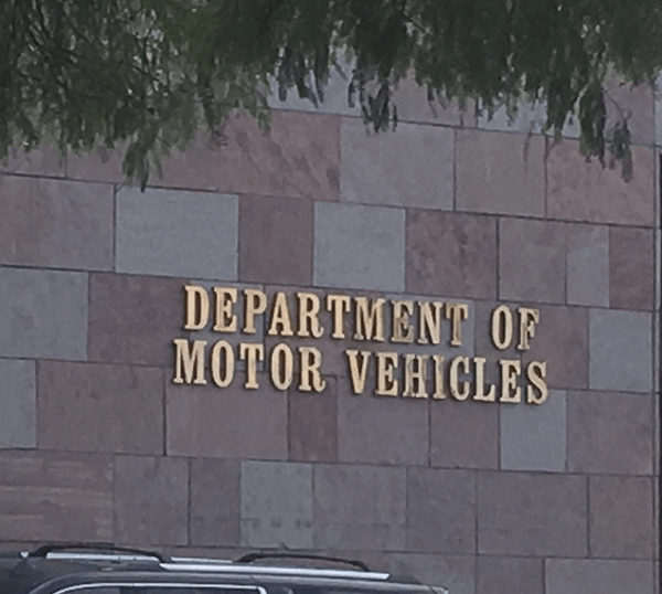 Photo of the exterior of a Department of Motor Vehicles (DMV) building. The wall is made of square and rectangular stone tiles in shades of beige and brown. Mounted in large gold letters is the text “DEPARTMENT OF MOTOR VEHICLES.” A portion of a parked vehicle and tree branches are visible in the foreground.