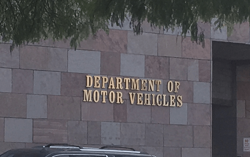Photo of the exterior of a Department of Motor Vehicles (DMV) building. The wall is made of square and rectangular stone tiles in shades of beige and brown. Mounted in large gold letters is the text “DEPARTMENT OF MOTOR VEHICLES.” A portion of a parked vehicle and tree branches are visible in the foreground.