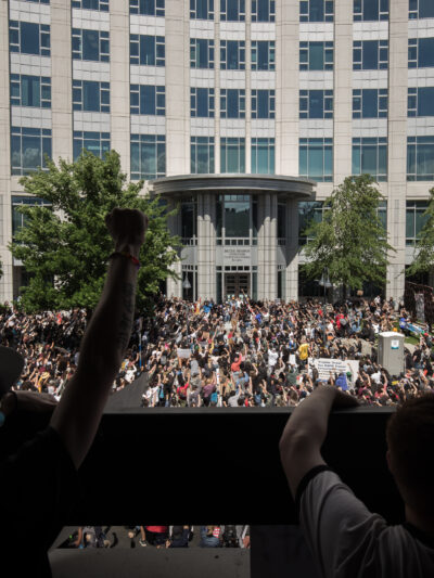 Large crowd gathered in front of a government building during a protest, with people raising their fists in solidarity. The photo is taken from behind a group of demonstrators on a balcony overlooking the scene, showing hundreds of participants filling the street below under a cloudy sky.