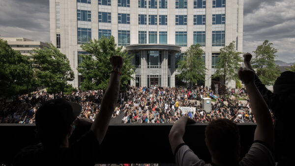 Large crowd gathered in front of a government building during a protest, with people raising their fists in solidarity. The photo is taken from behind a group of demonstrators on a balcony overlooking the scene, showing hundreds of participants filling the street below under a cloudy sky.