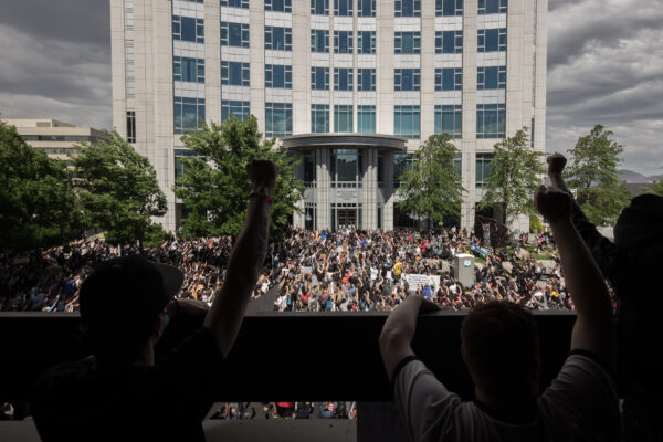 Large crowd gathered in front of a government building during a protest, with people raising their fists in solidarity. The photo is taken from behind a group of demonstrators on a balcony overlooking the scene, showing hundreds of participants filling the street below under a cloudy sky.