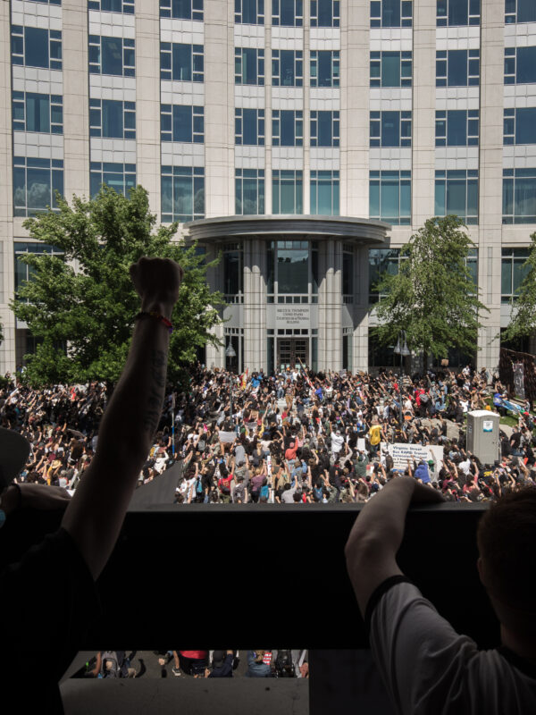 Large crowd gathered in front of a government building during a protest, with people raising their fists in solidarity. The photo is taken from behind a group of demonstrators on a balcony overlooking the scene, showing hundreds of participants filling the street below under a cloudy sky.
