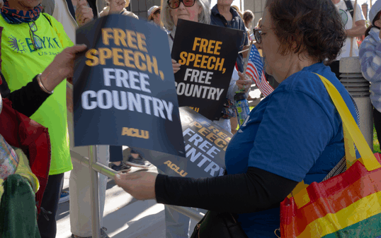 People gathered at a rally holding black ACLU signs that read ‘Free Speech, Free Country.’ A woman wearing a blue shirt and carrying a rainbow-striped tote bag hands out signs to others.