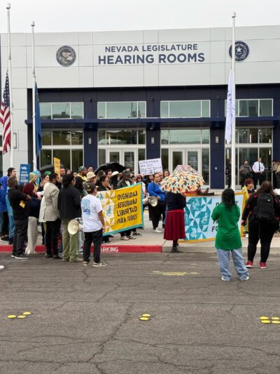 Group of people standing in front of Nevada Legislature Hearing Rooms with banners and signs.