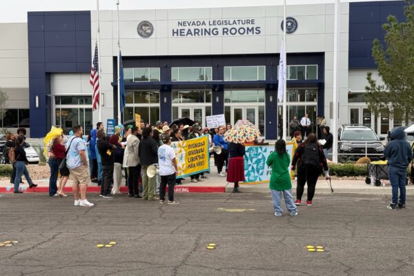 Group of people standing in front of Nevada Legislature Hearing Rooms with banners and signs.