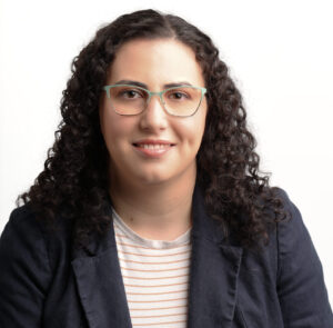 Headshot of Danielle. She is a white woman with medium length black curly hair. She is wearing glasses, a navy blazer, and a black and white striped shirt.