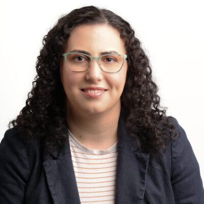 Headshot of Danielle. She is a white woman with medium length black curly hair. She is wearing glasses, a navy blazer, and a black and white striped shirt.