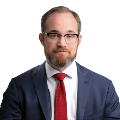 Headshot of Chris. He is a white man with glasses. He is wearing a navy blazer, white collared shirt, and a red tie.