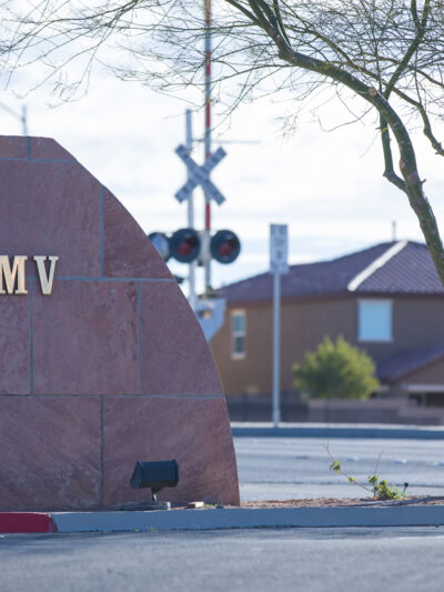 Stone monument sign reading ‘DMV’ positioned along a roadside curb. In the background are suburban houses, a railroad crossing signal, and a leafless tree.