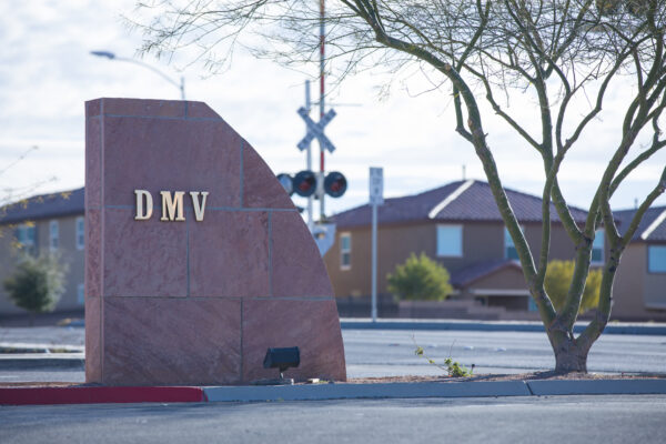 Stone monument sign reading ‘DMV’ positioned along a roadside curb. In the background are suburban houses, a railroad crossing signal, and a leafless tree.