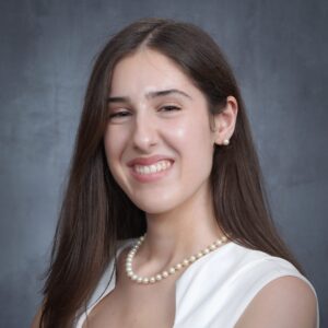 A smiling woman with long dark brown hair, wearing a white sleeveless top, pearl stud earrings, and a pearl necklace. The person faces the camera against a gray studio backdrop.