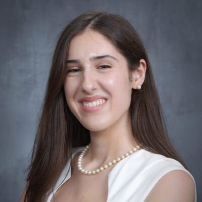 A smiling woman with long dark brown hair, wearing a white sleeveless top, pearl stud earrings, and a pearl necklace. The person faces the camera against a gray studio backdrop.