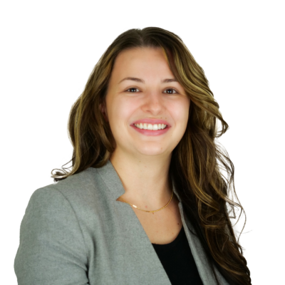 Woman with long curled brown hair, wearing a black top and grey blazer, smiling in front of a white background.