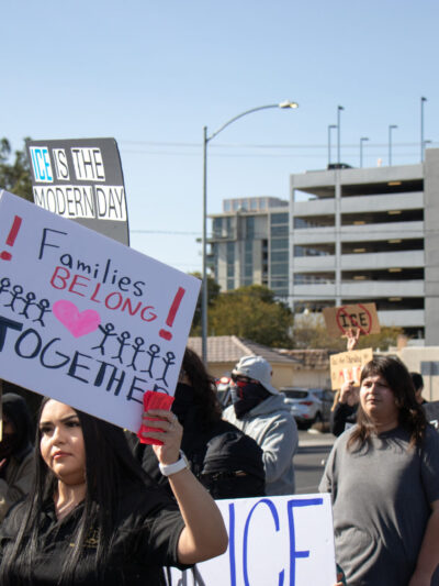 People march in a street protest carrying signs advocating for immigrant rights. In the foreground, a person holds a sign reading ‘Families Belong Together’ decorated with stick figures and a heart. Other visible signs include ‘ICE is the modern day…’ and ‘Democracy! Where the hell have you been, loca?!’ Participants walk through an urban area with office buildings, a parking garage, and traffic signs in the background.