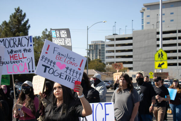 People march in a street protest carrying signs advocating for immigrant rights. In the foreground, a person holds a sign reading ‘Families Belong Together’ decorated with stick figures and a heart. Other visible signs include ‘ICE is the modern day…’ and ‘Democracy! Where the hell have you been, loca?!’ Participants walk through an urban area with office buildings, a parking garage, and traffic signs in the background.