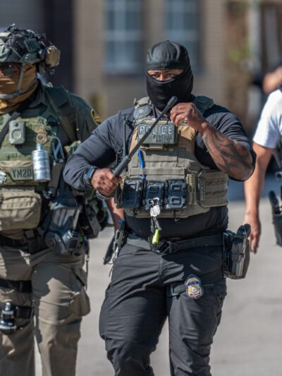 Several law enforcement officers wearing tactical gear, face coverings, and protective vests walk together in a parking lot. The officer in the center wears a vest labeled ‘Police’ and ‘Federal Agent’ and carries a baton, while others wear body armor, radios, and other equipment. Vehicles and buildings are visible in the background.