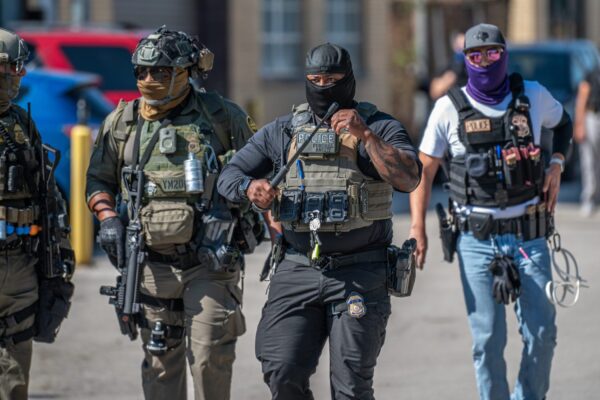 Several law enforcement officers wearing tactical gear, face coverings, and protective vests walk together in a parking lot. The officer in the center wears a vest labeled ‘Police’ and ‘Federal Agent’ and carries a baton, while others wear body armor, radios, and other equipment. Vehicles and buildings are visible in the background.