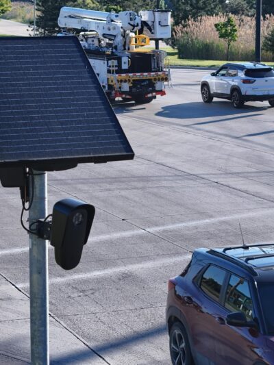 Flock camera mounted on a pole beside a busy multi-lane intersection. Several cars and a utility truck travel through the roadway while the camera faces passing traffic