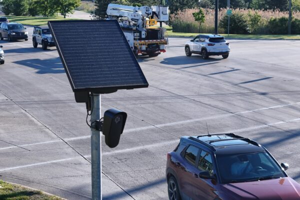 Flock camera mounted on a pole beside a busy multi-lane intersection. Several cars and a utility truck travel through the roadway while the camera faces passing traffic