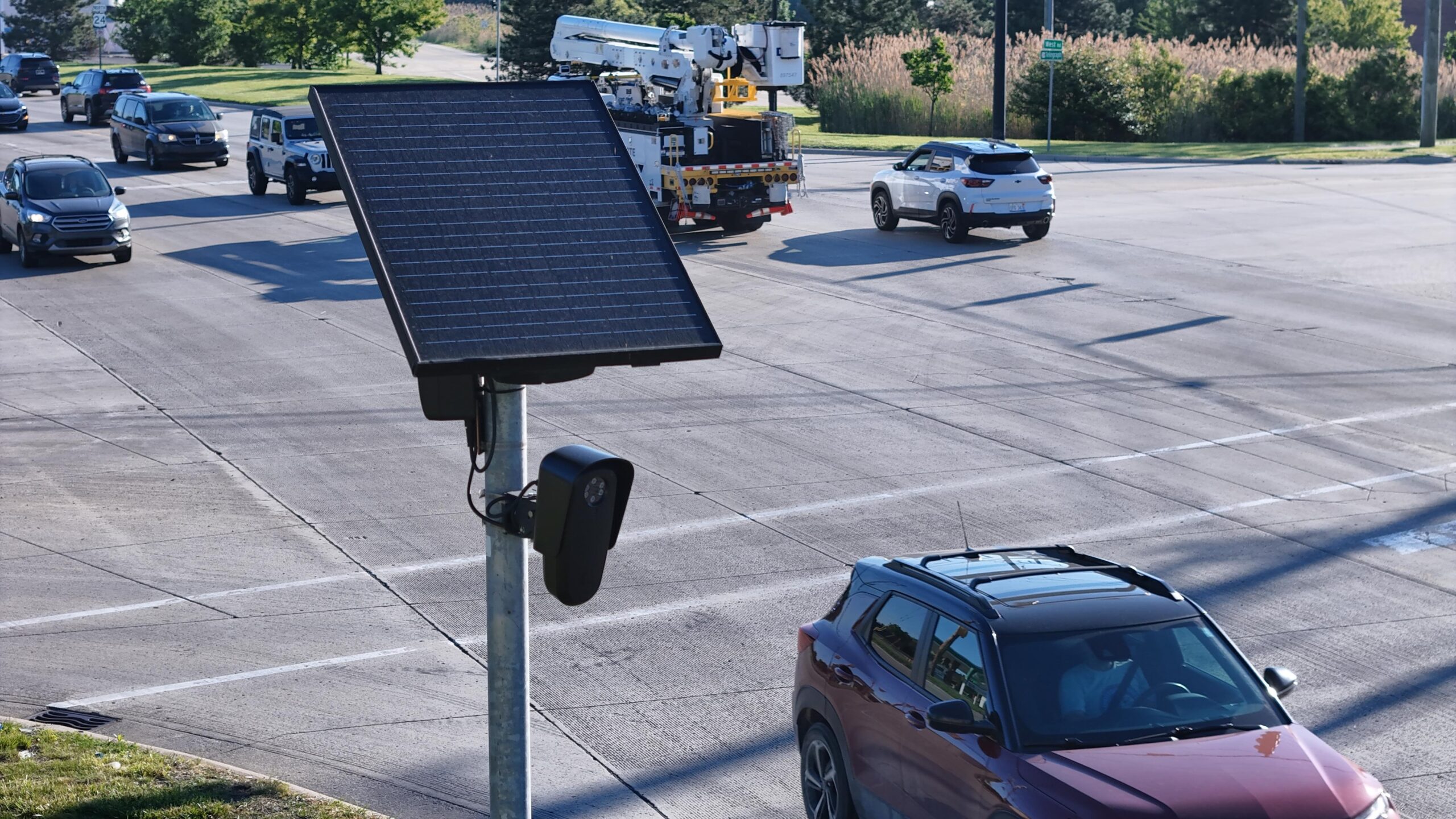 Flock camera mounted on a pole beside a busy multi-lane intersection. Several cars and a utility truck travel through the roadway while the camera faces passing traffic