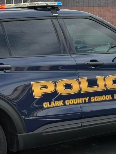 Close-up of a dark blue Clark County School District Police Department SUV with red and blue emergency lights illuminated. Large gold lettering on the side reads ‘Police’ and ‘Clark County School District,’ with a school building visible in the background.