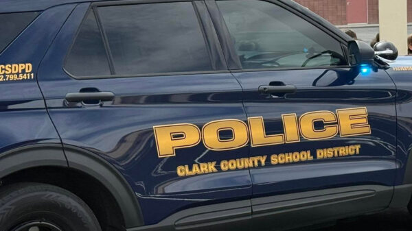 Close-up of a dark blue Clark County School District Police Department SUV with red and blue emergency lights illuminated. Large gold lettering on the side reads ‘Police’ and ‘Clark County School District,’ with a school building visible in the background.