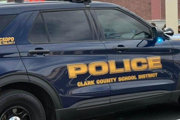 Close-up of a dark blue Clark County School District Police Department SUV with red and blue emergency lights illuminated. Large gold lettering on the side reads ‘Police’ and ‘Clark County School District,’ with a school building visible in the background.
