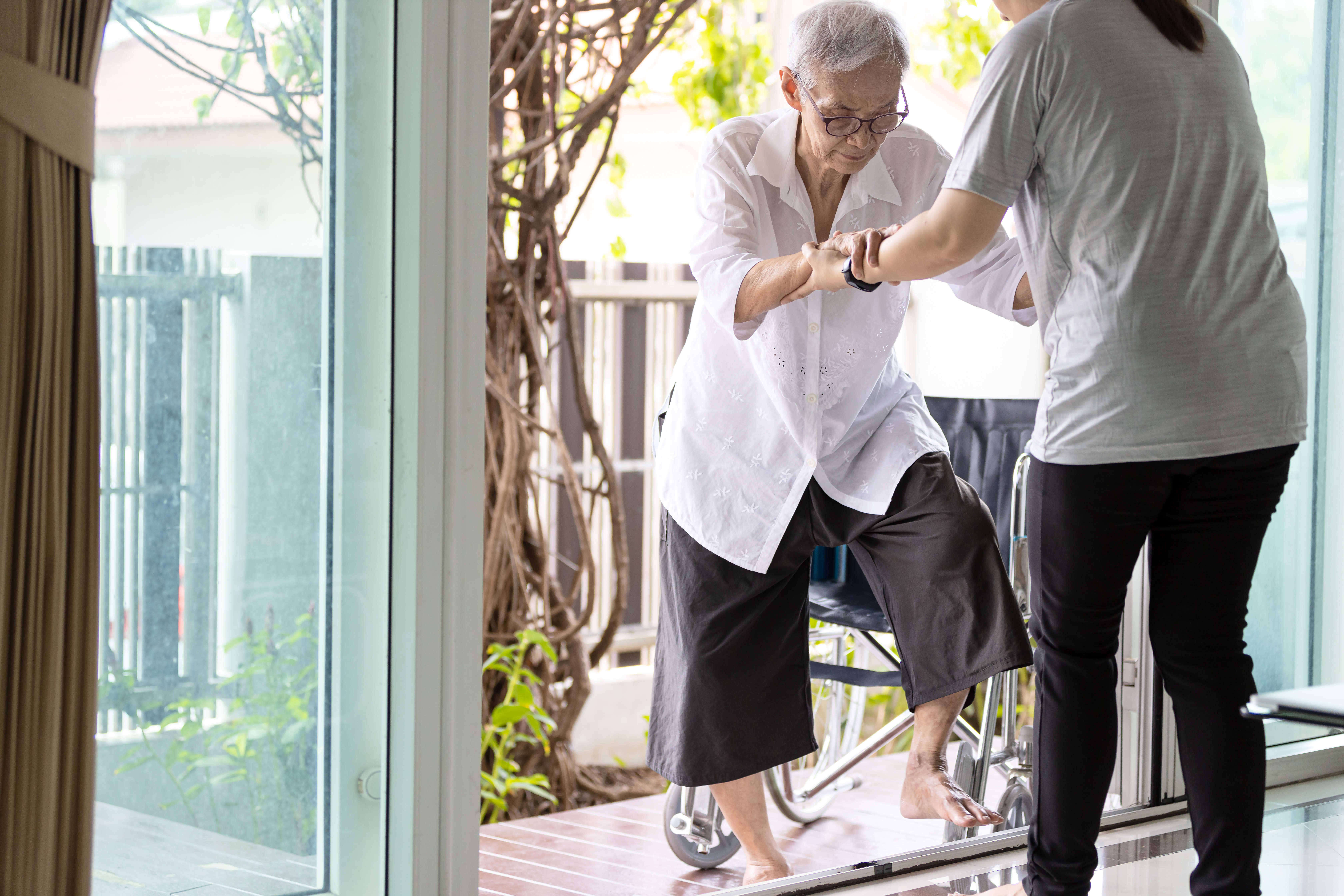 a woman helping an elderly women into the house. The doorway is a glass sliding door. 