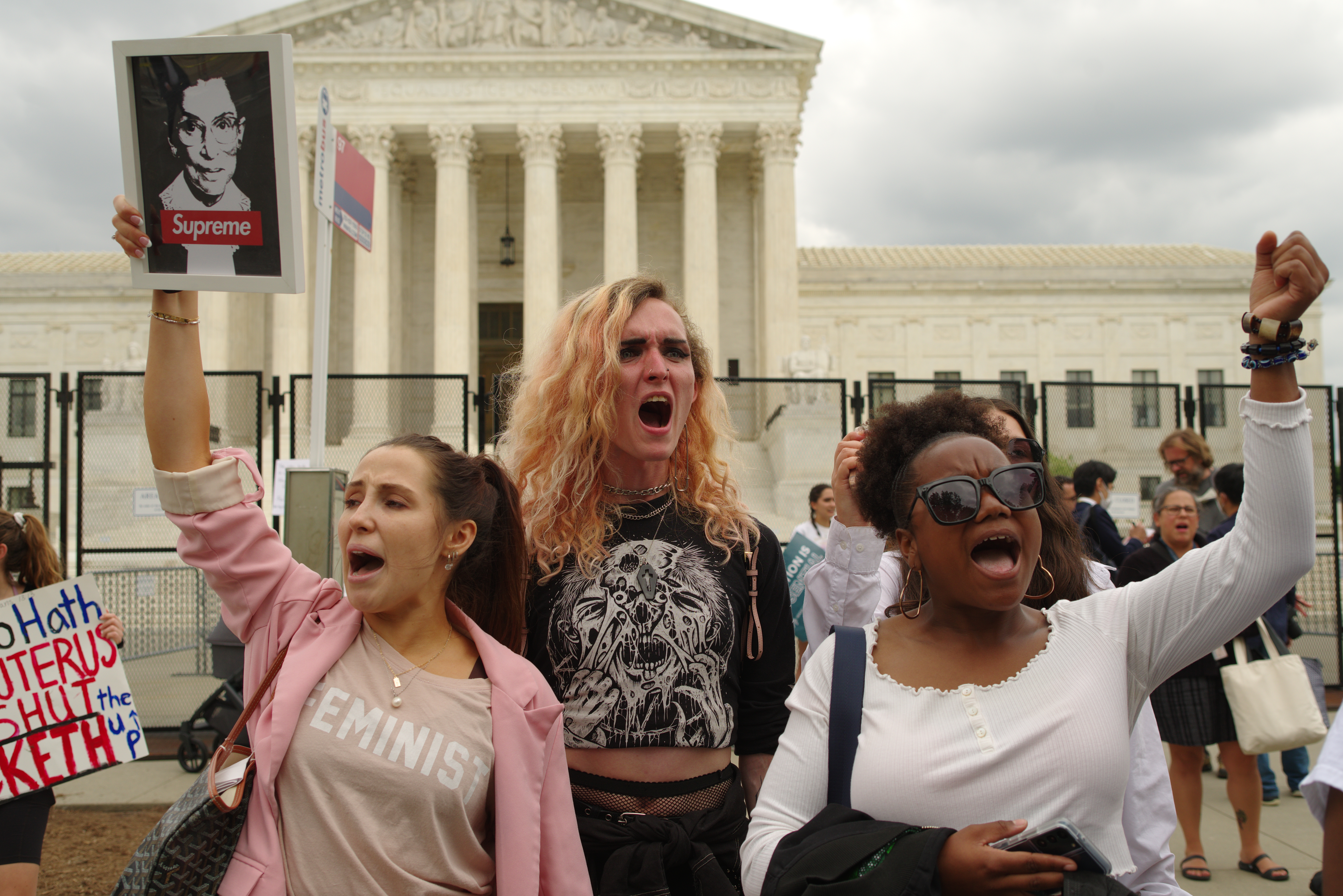 Three people in front of Supreme Court Building. Their fists are in the air and one is holding a picture of RBG.