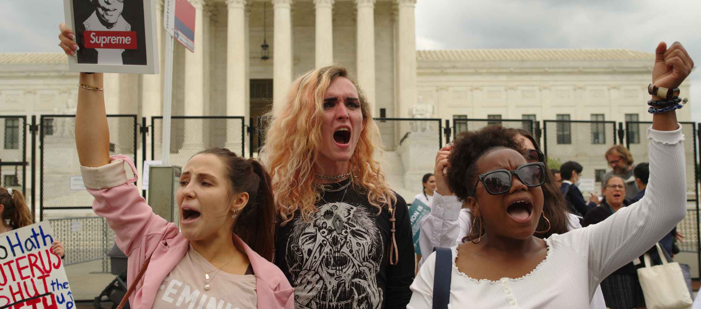 Three people in front of Supreme Court Building. Their fists are in the air and one is holding a picture of RBG.