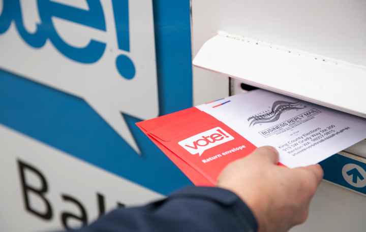 A hand holding a ballot that is half way in the slot of a ballot box.