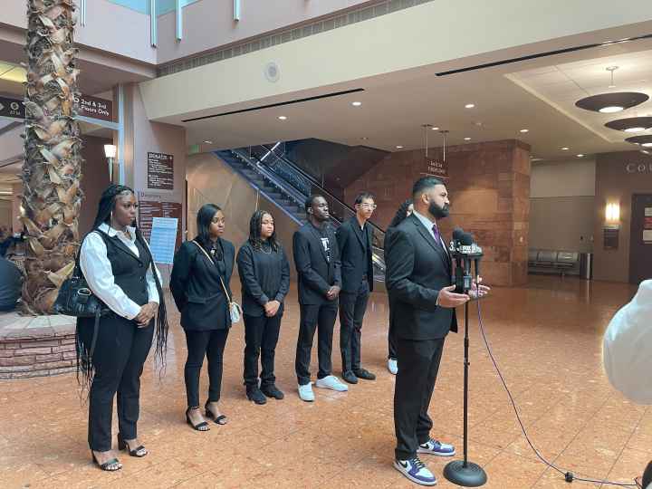 Athar Haseebullah standing at a microphone speaking to the press while the 6 Emerging Leaders Board Members stand behind him in a line.