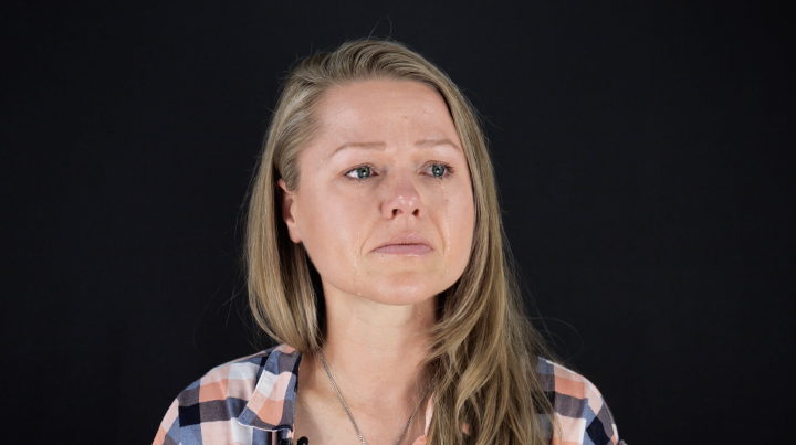 Client, Rebecca Leavitt. She is a white woman with blonde hair, blue eyes, and is wearing a blue, pink, and white checkered collared shirt. She is looking off to the side and has tears down her face.