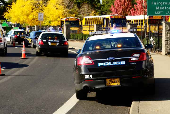 Line of police cars parked in front of school. There is also a line of school buses.