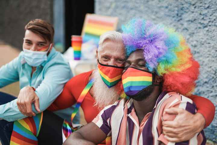 Three men at a LGBTQ+ parade. They are sitting on the curb. The man on the right is a black man with a rainbow afro wig on and a rainbow mask. He is being hugged by the man to his left who also has a rainbow mask on. The third man has a blue mask on.