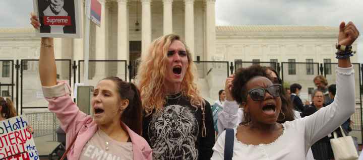 Three people in front of Supreme Court Building. Their fists are in the air and one is holding a picture of RBG.