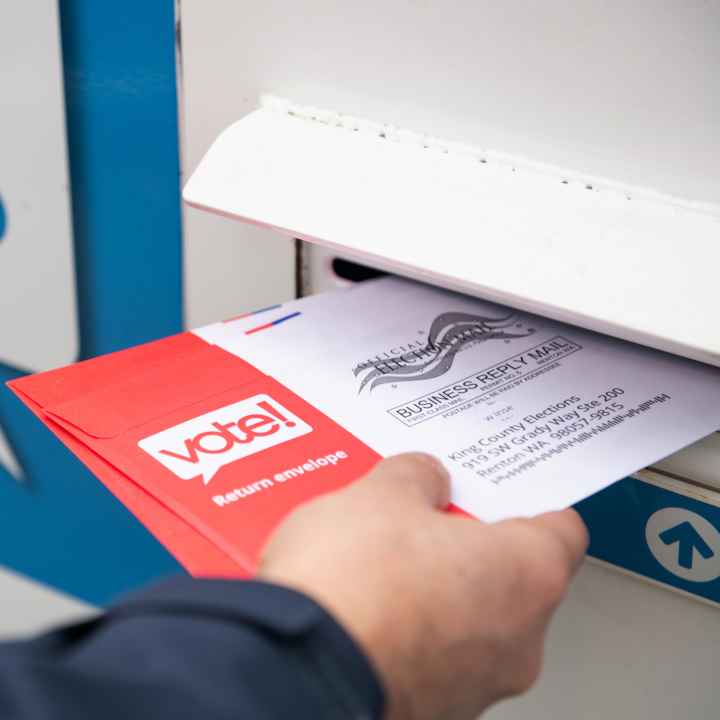 A hand holding a ballot that is half way in the slot of a ballot box.