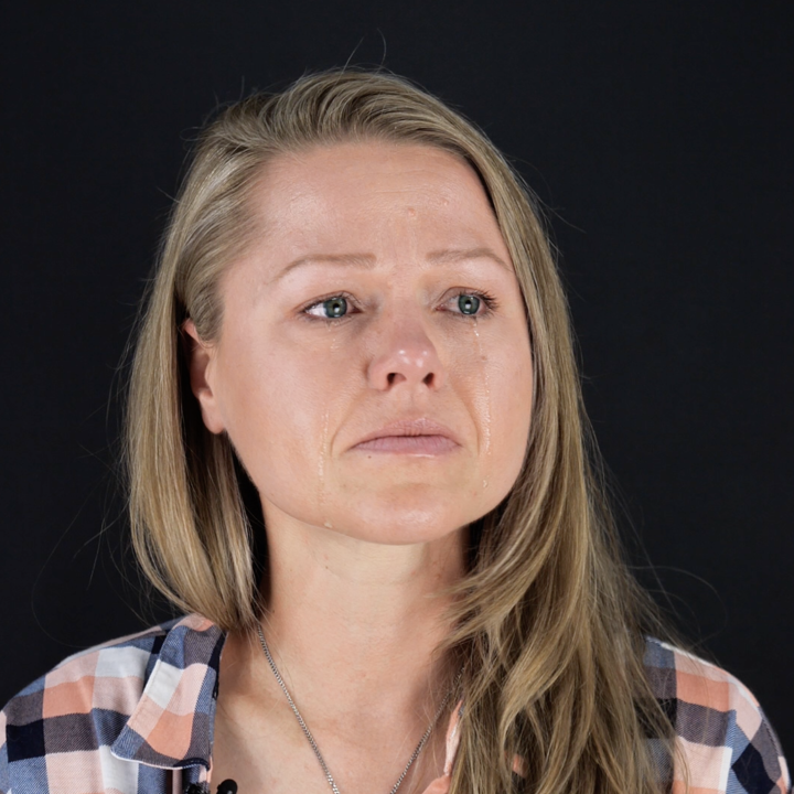Client, Rebecca Leavitt. She is a white woman with blonde hair, blue eyes, and is wearing a blue, pink, and white checkered collared shirt. She is looking off to the side and has tears down her face.