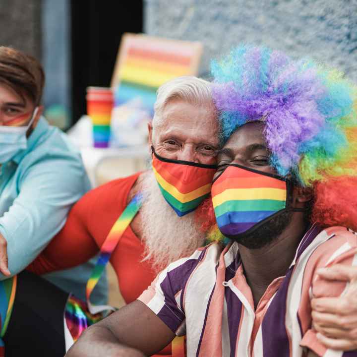 Three men at a LGBTQ+ parade. They are sitting on the curb. The man on the right is a black man with a rainbow afro wig on and a rainbow mask. He is being hugged by the man to his left who also has a rainbow mask on. The third man has a blue mask on.