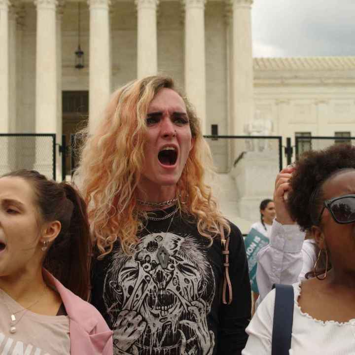 Three people in front of Supreme Court Building. Their fists are in the air and one is holding a picture of RBG.